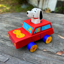 Toy car with a dog figure on top, resembling Snoopy, on a wooden surface.