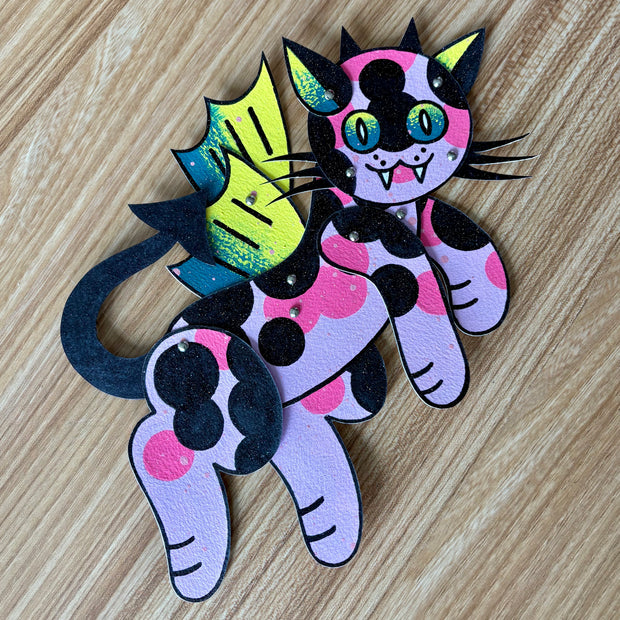 Paper sculpture assembled with silver brads of a pink cat with black and bright pink spots. It has a set of neon green and blue dragon-like wings and horns. 