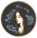 Illustration on round wooden panel of a girl, visible from the neck up, looking downward with a weepy expression. She has long black hair that blends into the black background and flowers made of abalone surround her.