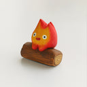 A small flame-shaped figurine with a red-to-orange gradient face and white eyes, sitting on a brown wooden log base against a plain light background.