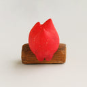 A small flame-shaped figurine with a red-to-orange gradient face and white eyes, sitting on a brown wooden log base against a plain light background.