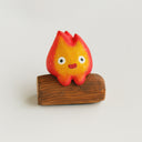 A small flame-shaped figurine with a red-to-orange gradient face and white eyes, sitting on a brown wooden log base against a plain light background.
