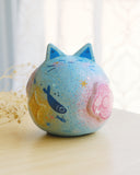 Sculpture of a round, cat with pointed ears, a small smiling face and painted fish on its body. There are raised elements on its body, such as flowers and stars.