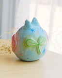 Sculpture of a round, cat with pointed ears, a small smiling face and painted fish on its body. There are raised elements on its body, such as flowers and stars.