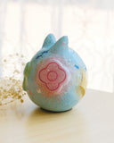 Sculpture of a round, cat with pointed ears, a small smiling face and painted fish on its body. There are raised elements on its body, such as flowers and stars.