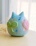 Sculpture of a round, cat with pointed ears, a small smiling face and painted fish on its body. There are raised elements on its body, such as flowers and stars.