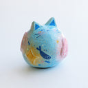 Sculpture of a round, cat with pointed ears, a small smiling face and painted fish on its body. There are raised elements on its body, such as flowers and stars.