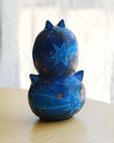 Sculpture of 2 cat heads, stacked atop of one another and painted to look like a night sky with shooting stars. Each head has a tiny, cute cat face and gold sparkly stars.