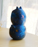 Sculpture of 2 cat heads, stacked atop of one another and painted to look like a night sky with shooting stars. Each head has a tiny, cute cat face and gold sparkly stars.