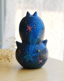 Sculpture of 2 cat heads, stacked atop of one another and painted to look like a night sky with shooting stars. Each head has a tiny, cute cat face and gold sparkly stars.