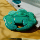 A painted clay sculpture of a sleeping dinosaur, curled into itself like a sleeping cat. It is a teal with stars painted and glitter stars pasted on.
