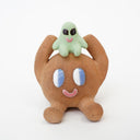 Sculpture of a brown, rounded character with legs sitting down. It holds a green smiling alien character atop its head.
