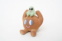 Sculpture of a brown, rounded character with legs sitting down. It holds a green smiling alien character atop its head.