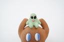 Sculpture of a brown, rounded character with legs sitting down. It holds a green smiling alien character atop its head.