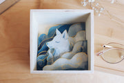 Diorama style artwork of a blue wolf, seen only from the chest up, looking off to the left. It is surrounded by blue watercolor clouds with subtle gold outlining.  Artwork is contained in an open faced wooden box.
