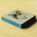 Painting on exposed wooden panel of a calico cat's head, floating and wearing a blue collar with a small gold bell. It has one blue eye and one brown eye, and looks upward at a tiny blue fish.