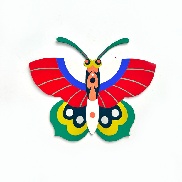 Die cut, brightly painted wooden sculpture of a butterfly with multicolored wings. Top wings are white, blue and red and bottom set are green, yellow and navy blue. It has a patterned body and green antennae. 