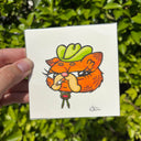 A hand holds a square white artwork featuring a cartoon orange cat wearing a green cowboy hat and holding a cigarette, with a blurred green leafy background behind it.