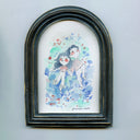 Stylistically messy watercolor painting of a couple sitting in a blurry field of flowers and plants. They hold hands and look up towards the sky. Piece is in an arch shaped frame.