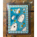 A whimsical illustration of white ghosts floating on a bright blue background with small yellow stars, sealed in a clear plastic sleeve and placed on a wooden surface.