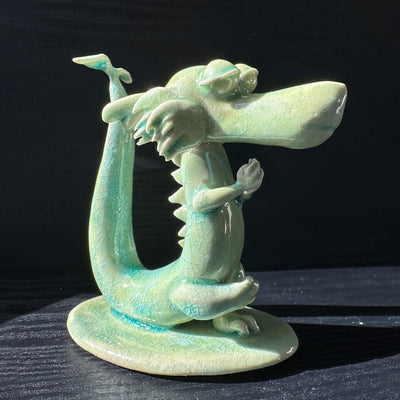 Jade colored ceramic sculpture of a dragon with a large front snout, positioned in a yoga pose with one leg up and its hands at its chest.