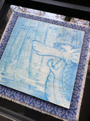 Illustration done in blue on wood board of a dragon with a long snout, holding its hands together in a bamboo forest with a pond. On a blue cloth and within a clear frame.