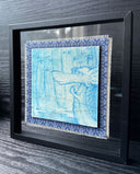 Illustration done in blue on wood board of a dragon with a long snout, holding its hands together in a bamboo forest with a pond. On a blue cloth and within a clear frame.