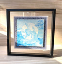 Illustration done in blue on wood board of a dragon with a long snout, doing a yoga stretch in front of a large full moon. On a blue cloth and within a clear frame.
