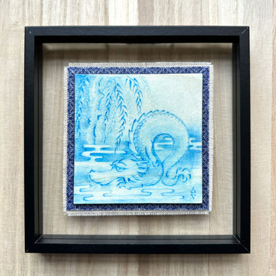 Illustration done in blue on wood board of a dragon with a long snout, doing a yoga stretch under a willow like tree. Wood board is on a blue cloth and within a clear frame.