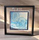 Illustration done in blue on wood board of a dragon with a long snout, doing a yoga stretch under a willow like tree. Wood board is on a blue cloth and within a clear frame.