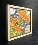 Painted ceramic tile with bright green and orange coloring with 3 characters surrounding the exterior. One is Totoro with a leaf umbrella, the other is a dark blue smaller Totoro and the last is a small white Totoro.