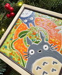 Painted ceramic tile with bright green and orange coloring with 3 characters surrounding the exterior. One is Totoro with a leaf umbrella, the other is a dark blue smaller Totoro and the last is a small white Totoro.