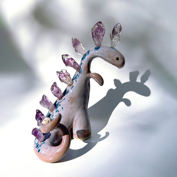 Ceramic sculpture of a purple dinosaur with a curling tail and amethyst crystals coming out of its back, like spikes. On its tail is a tiny character, clinging on.