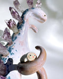Ceramic sculpture of a purple dinosaur with a curling tail and amethyst crystals coming out of its back, like spikes. On its tail is a tiny character, clinging on.
