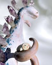 Ceramic sculpture of a purple dinosaur with a curling tail and amethyst crystals coming out of its back, like spikes. On its tail is a tiny character, clinging on.