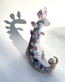 Ceramic sculpture of a purple dinosaur with a curling tail and amethyst crystals coming out of its back, like spikes. On its tail is a tiny character, clinging on.