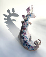 Ceramic sculpture of a purple dinosaur with a curling tail and amethyst crystals coming out of its back, like spikes. On its tail is a tiny character, clinging on.