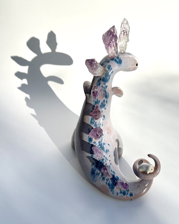 Ceramic sculpture of a purple dinosaur with a curling tail and amethyst crystals coming out of its back, like spikes. On its tail is a tiny character, clinging on.