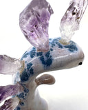 Ceramic sculpture of a purple dinosaur with a curling tail and amethyst crystals coming out of its back, like spikes. On its tail is a tiny character, clinging on.