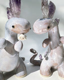 Ceramic sculpture of a purple dinosaur with a curling tail and amethyst crystals coming out of its back, like spikes. On its tail is a tiny character, clinging on.