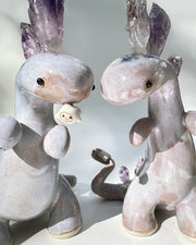 Ceramic sculpture of a purple dinosaur with a curling tail and amethyst crystals coming out of its back, like spikes. On its tail is a tiny character, clinging on.