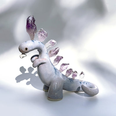 Ceramic sculpture of a purple dinosaur with a curling tail and amethyst crystals coming out of its back, like spikes. Dangling from its mouth is a tiny character.
