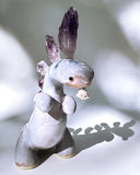 Ceramic sculpture of a purple dinosaur with a curling tail and amethyst crystals coming out of its back, like spikes. Dangling from its mouth is a tiny character.