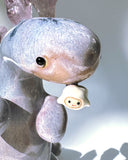 Ceramic sculpture of a purple dinosaur with a curling tail and amethyst crystals coming out of its back, like spikes. Dangling from its mouth is a tiny character.