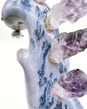 Ceramic sculpture of a purple dinosaur with a curling tail and amethyst crystals coming out of its back, like spikes. Dangling from its mouth is a tiny character.