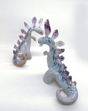 Ceramic sculpture of a purple dinosaur with a curling tail and amethyst crystals coming out of its back, like spikes. Dangling from its mouth is a tiny character.