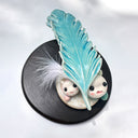 Ceramic sculpture of 2 small, cute monsters holding up a blue ceramic feather, as if gifting it to the viewer. Attached the the ceramic feather is a real white feather.