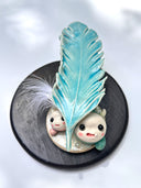 Ceramic sculpture of 2 small, cute monsters holding up a blue ceramic feather, as if gifting it to the viewer. Attached the the ceramic feather is a real white feather.