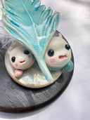 Ceramic sculpture of 2 small, cute monsters holding up a blue ceramic feather, as if gifting it to the viewer. Attached the the ceramic feather is a real white feather.