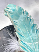 Ceramic sculpture of 2 small, cute monsters holding up a blue ceramic feather, as if gifting it to the viewer. Attached the the ceramic feather is a real white feather.